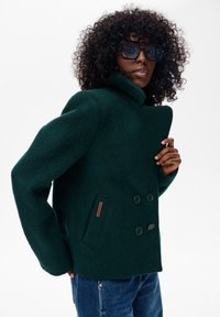 Kobieta z kręconymi włosami, nosząca ciemne okulary przeciwsłoneczne, ciemnozielony, dwurzędowy wełniany płaszcz oraz niebieskie dżinsy na białym tle.