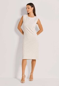 Knielanges, ärmelloses Kleid aus strukturiertem weißen Stoff mit rundem Ausschnitt. Kombiniert mit nude-farbenen, hochhackigen Sandalen.