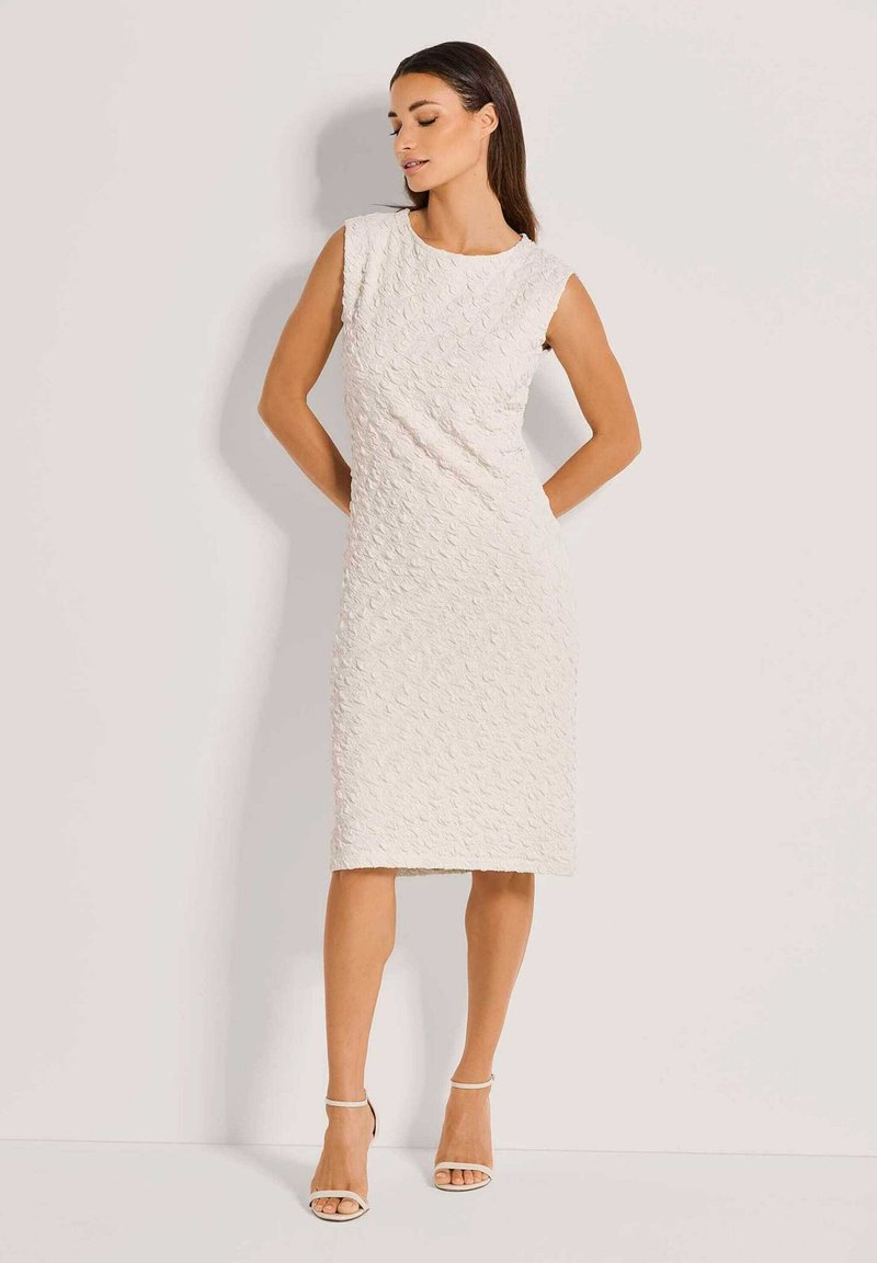 Knielanges, ärmelloses Kleid aus strukturiertem weißen Stoff mit rundem Ausschnitt. Kombiniert mit nude-farbenen, hochhackigen Sandalen.