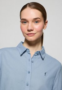 Camisa de botón de color azul claro en una tela suave, con cuello y un pequeño diseño bordado en el lado izquierdo del pecho.