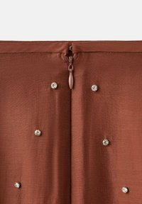 Braunes Stoffkleidungsstück mit einem zentralen Reißverschluss und mehreren verstreuten, glänzenden Strass-Verzierungen auf der Oberfläche.