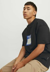 Sort bomulds t-shirt med et blåt og hvidt geometrisk logo patch på brystet. Modellen bærer khaki shorts og sidder mod en ensfarvet baggrund.