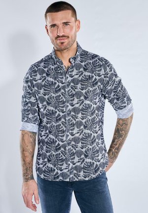 Mann mit kurzen Haaren und Tätowierungen, trägt ein gemustertes schwarz-weißes Hemd mit Knöpfen und hochgekrempelten Ärmeln sowie dunkelblaue Jeans, stehend.