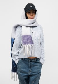 Χοντρό πλεκτό πουλόβερ γαλάζιου χρώματος, συνδυασμένο με κασκόλ με διαβάθμιση γκρι και μοβ με φούντες. Μαύρο καπέλο μπέιζμπολ ολοκληρώνει την εμφάνιση.