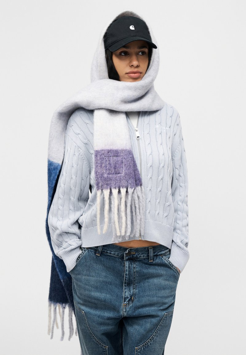 Pull bleu clair en grosse maille torsadée, associé à une écharpe dégradée grise et violette à franges. Une casquette de baseball noire complète la tenue.