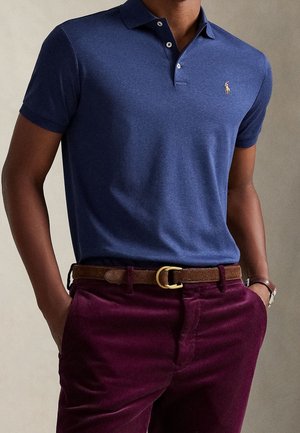 Mężczyzna w granatowej koszulce polo z małym logo, burgundowych spodniach ze sztruksu, brązowym pasku i zegarku na nadgarstku, z rękami w kieszeniach.