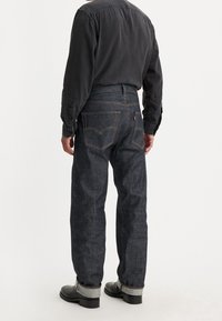 Mörkblå denimjeans med rak skärning, kontrasterande gul söm och bakfickor med en distinkt logotyp. Bärs med svarta stövlar.
