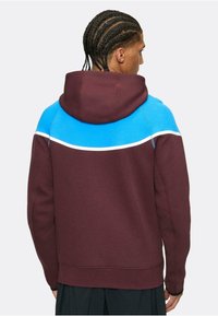 Sudadera burdeos con una sección azul en el hombro y una franja blanca de acento. Presenta un estilo de pullover con capucha y puños acanalados.
