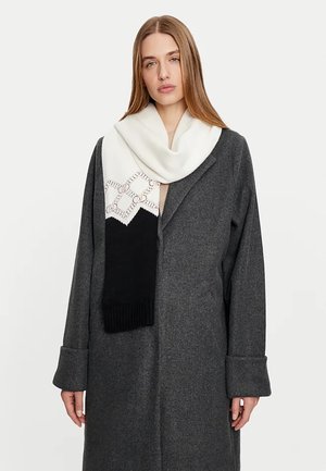 Sciarpa bianca e nera con un design geometrico, realizzata in tessuto morbido, con una texture a costine alle estremità, drappeggiata su un cappotto grigio.