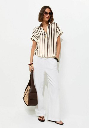 Vrouw met oversized zonnebril, gestreept T-shirt met korte mouwen, witte broek met wijde pijpen, houdt een tweekleurige bruine tas en zwarte slippers vast.