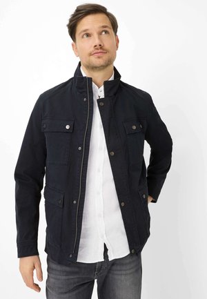 Mann trägt eine offene schwarze Jacke über einem weißen Hemd mit Knöpfen und grauen Jeans, schaut leicht nach oben und hat eine Hand in der Hüfte.