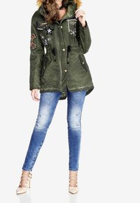 Veste parka verte avec capuche en fourrure, ornée de patchs brodés et de boutons ; associée à un jeans skinny bleu clair et des talons beiges.