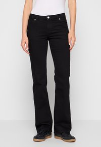Femme portant un jean bootcut noir, des baskets noires à lacets et un haut blanc sans manches sur un fond clair uni.
