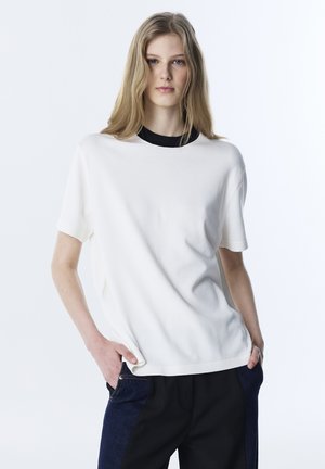 Junge Frau mit blondem Haar, die ein weißes Kurzarmoberteil über einem schwarzen Shirt und blau-schwarz farblich abgesetzte Hosen trägt, mit den Händen in den Taschen.