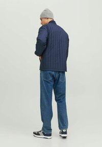 Giacca trapuntata blu navy con colletto alto, indossata su jeans di denim. Accompagnata da un berretto grigio e scarpe nere con dettagli grigi.
