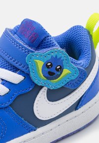 Niebieskie trampki z designem z skóry i siateczki, białym logo Nike, oraz naklejką z rysunkowym niebieskim charakterem na pasku. Żółta pętla na pięcie.