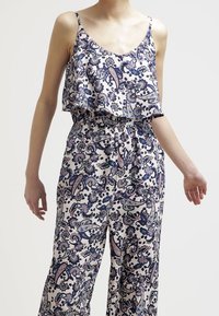 Kobieta w bezrękawowym, luźnym kombinezonie w kwiaty z niebieskimi, fioletowymi i beżowymi wzorami paisley, stojąca na jednolitym tle.