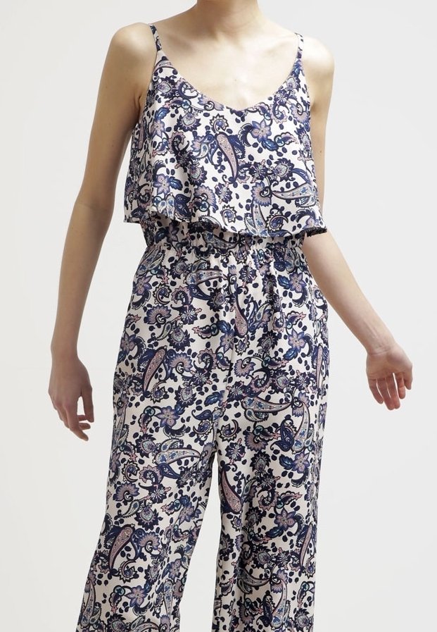 Kobieta w bezrękawowym, luźnym kombinezonie w kwiaty z niebieskimi, fioletowymi i beżowymi wzorami paisley, stojąca na jednolitym tle.
