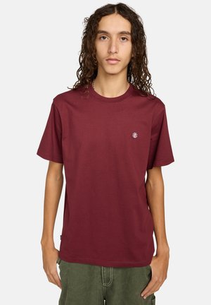 T-shirt en coton bordeaux, col rond, manches courtes, avec un petit logo circulaire sur le côté gauche de la poitrine. Texture lisse, coupe décontractée.