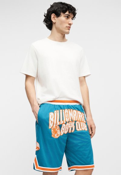 Fiatal férfi egyszerű fehér pólóban és kék kosárlabda nadrágban, amelyen fehér és narancssárga felirat olvasható: "Billionaire Boys Club".