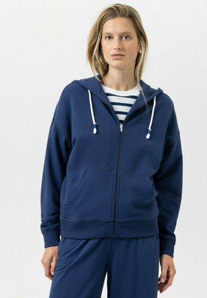 Femme portant un sweat à capuche zippé bleu marine sur un pantalon bleu marine et une chemise blanche à rayures horizontales noires, debout devant un fond uni.