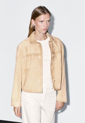 Tan suede jack met een kraag, voorzijde met knopen en zijzakken. Draagt over een wit T-shirt en gecombineerd met witte broek.