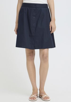 Kvinde iført navyblåt A-line nederdel med knapper foran kombineret med hvide flade sandaler med stropper mod en ensfarvet baggrund.