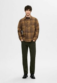 Brązowa i czarna koszula w kratę z guzikami, z dwiema kieszeniami na piersiach, połączona z ciemnozielonymi spodniami i czarnymi butami. Prosty, luźny krój.