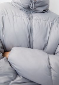 Chaqueta acolchada de color gris claro con cuello alto, cremallera frontal completa y textura acolchada. El material parece suave e insulated para mantener el calor.