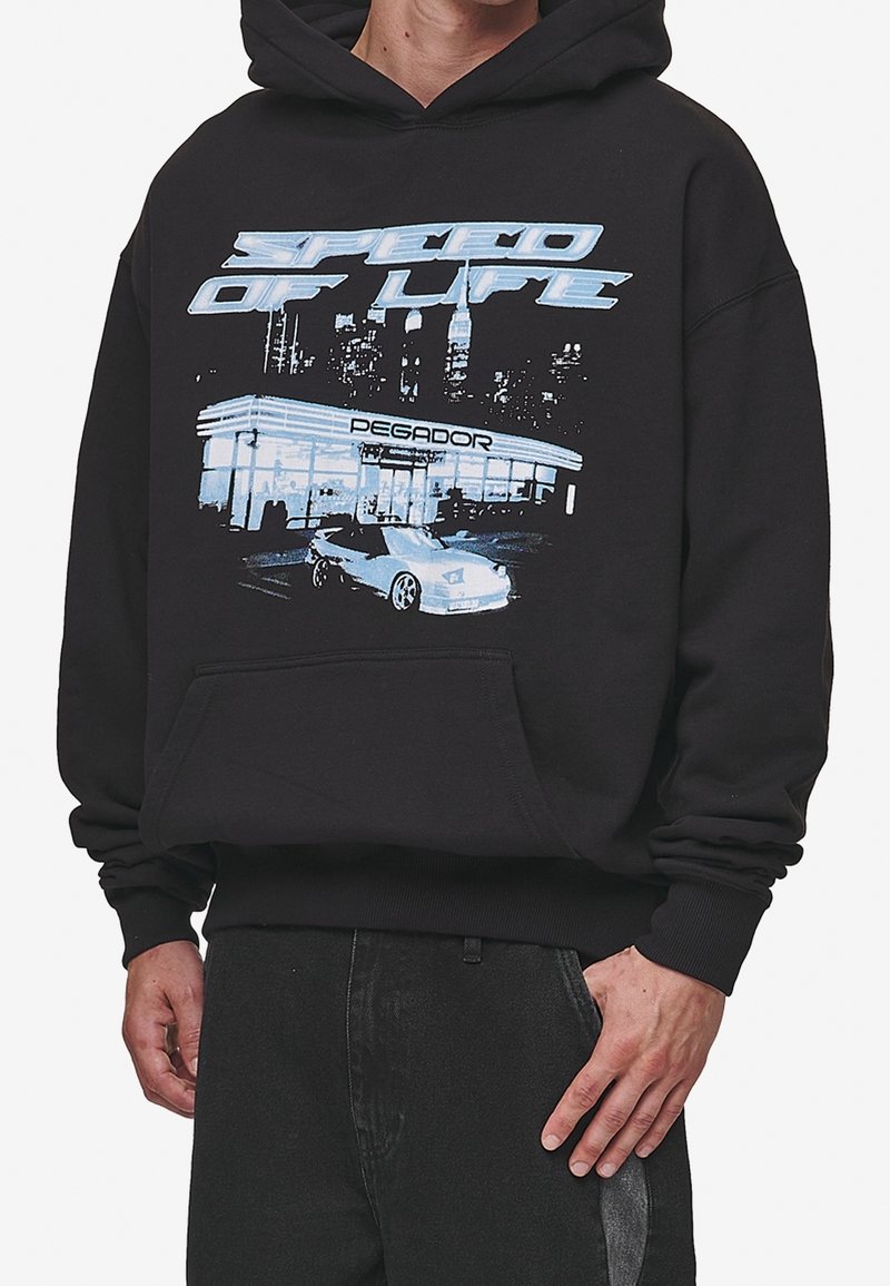 Svart hoodie med en grafisk design som har texten "LIVETS HASTIGHET" och en stadsbild, som visar en bil och en bensinstation i ljusblå accenter.