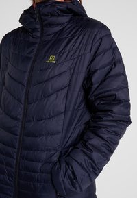 Navy Pufferjacke mit hohem Kragen, diagonal gestepptem Muster, Reißverschluss und kleinem grünen Logo auf der Brust.