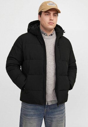 Homme portant une casquette beige, une doudoune noire à capuche, un pull gris, une chemise à carreaux et un jean bleu, debout les mains dans les poches de la veste.