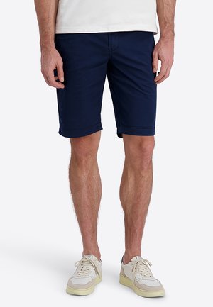 Mann trägt marineblaue knielange Shorts, weiße Sneakers und ein weißes Hemd und steht vor einem einfarbigen hellen Hintergrund.