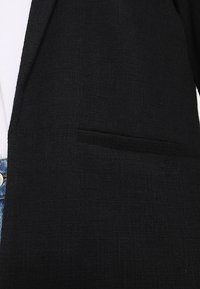 Černý blejzr s texturovanou látkou, vybavený jedním kapsou na levé straně, střih na míru a viditelné okraje klopy.