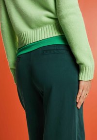 Světle zelený pletený svetr s žebrovanými manžetami, přes tmavě zelený top, doplněný o tmavě zelené kalhoty s zadními kapsami. Pozadí je oranžové.