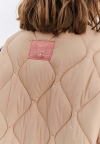 Prošívaná vestuška světle béžové barvy s texturovaným povrchem, s růžovou etiketou se textem a ozdobným prošíváním.