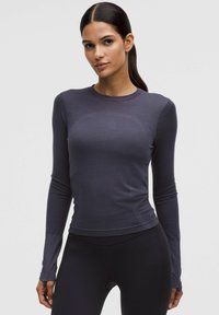 Tumedate juustega naine, kellel on madal ponisaba, kannab sobivat tumeda halli pikkade varrukatega topi ja musti legginsid, seistes lihtsa heleda tausta ees.