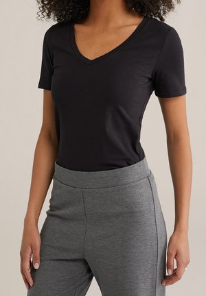 Kobieta w prostym czarnym T-shircie z dekoltem w serek i krótkim rękawem, wpuszczonym w wysoki stan szarych spodni z bocznymi szwami.