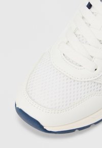 Λευκό αθλητικό παπούτσι με πάνω μέρος από διχτυωτό υλικό, με λείες δερμάτινες λεπτομέρειες και μπλε λαστιχένια σόλα. Τα κορδόνια είναι επίπεδα και υφασμένα.