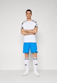 Hvid atletisk skjorte med sorte striber, blå shorts, hvide sokker med sorte striber og hvide støvler, stående med armene korslagt.