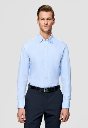 Mężczyzna w jasnoniebieskiej koszuli zapinanej na guziki, włożonej w czarne spodnie z czarnym paskiem, stojący na jednolitym tle.