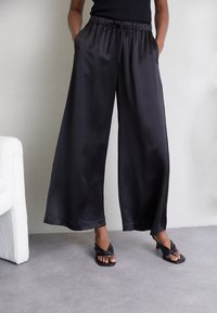 Svarta satinbyxor med vida ben, elastisk midja, snördetalj, sidofickor och en mjuk textur. Bärs med svarta slidesandaler.