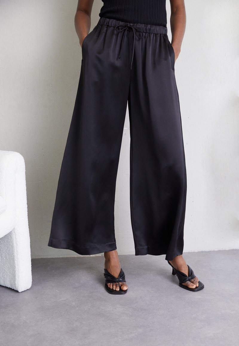 Svarta satinbyxor med vida ben, elastisk midja, snördetalj, sidofickor och en mjuk textur. Bärs med svarta slidesandaler.
