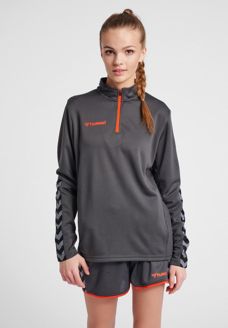 Donkergrijze sporttop met kwart rits, voorzien van oranje accenten en logo, met een gestructureerde stof en mouwdetails in houndstooth-patroon.