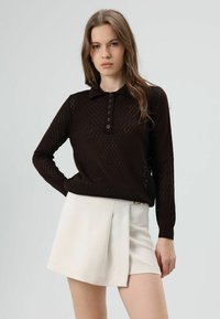 Bruine gebreide trui met een ruitpatroon, een kraag en vijf knopen aan de voorkant, gecombineerd met een geplooide witte rok.