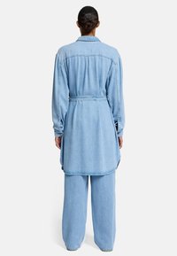 Niebieska sukienka koszulowa z dżinsu z paskiem w talii, długimi rękawami i zaokrąglonym dołem, w komplecie z szerokimi spodniami pasującymi do całości.