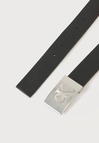 Μαύρη δερμάτινη ζώνη με ασημί ορθογώνια αγκράφα που φέρει ανάγλυφο λογότυπο «CK», απλωμένη σε λευκή επιφάνεια.