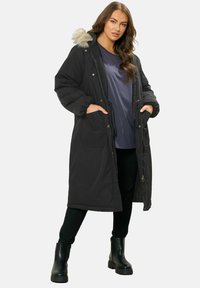 Musta eristetty parka, jossa on tekoturkiksella vuorattu huppu, napilla suljettava etuosa, kaksi sivutaskua ja ribattu hihansuu. Päällä sininen paita ja mustat housut.