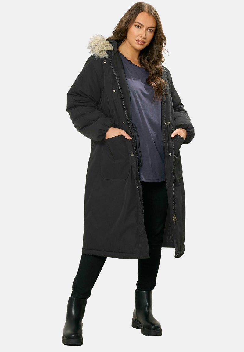 Musta eristetty parka, jossa on tekoturkiksella vuorattu huppu, napilla suljettava etuosa, kaksi sivutaskua ja ribattu hihansuu. Päällä sininen paita ja mustat housut.