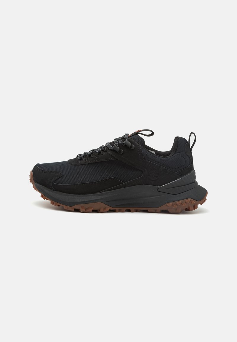 Chaussure de sport noire avec une tige en mesh et en daim, semelle extérieure en caoutchouc, panneaux texturés et crampons de traction bruns contrastants. Elle dispose d'accents réfléchissants.